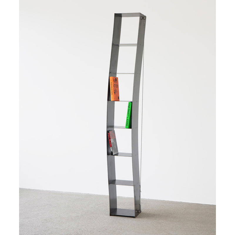 A curved metal bookshelf from Moormann, the Gespanntes Regal, stands tall against a white wall on a gray floor. Supported by steel cables, it displays an orange book at the top shelf, a green book in the middle, and a red one at the bottom.