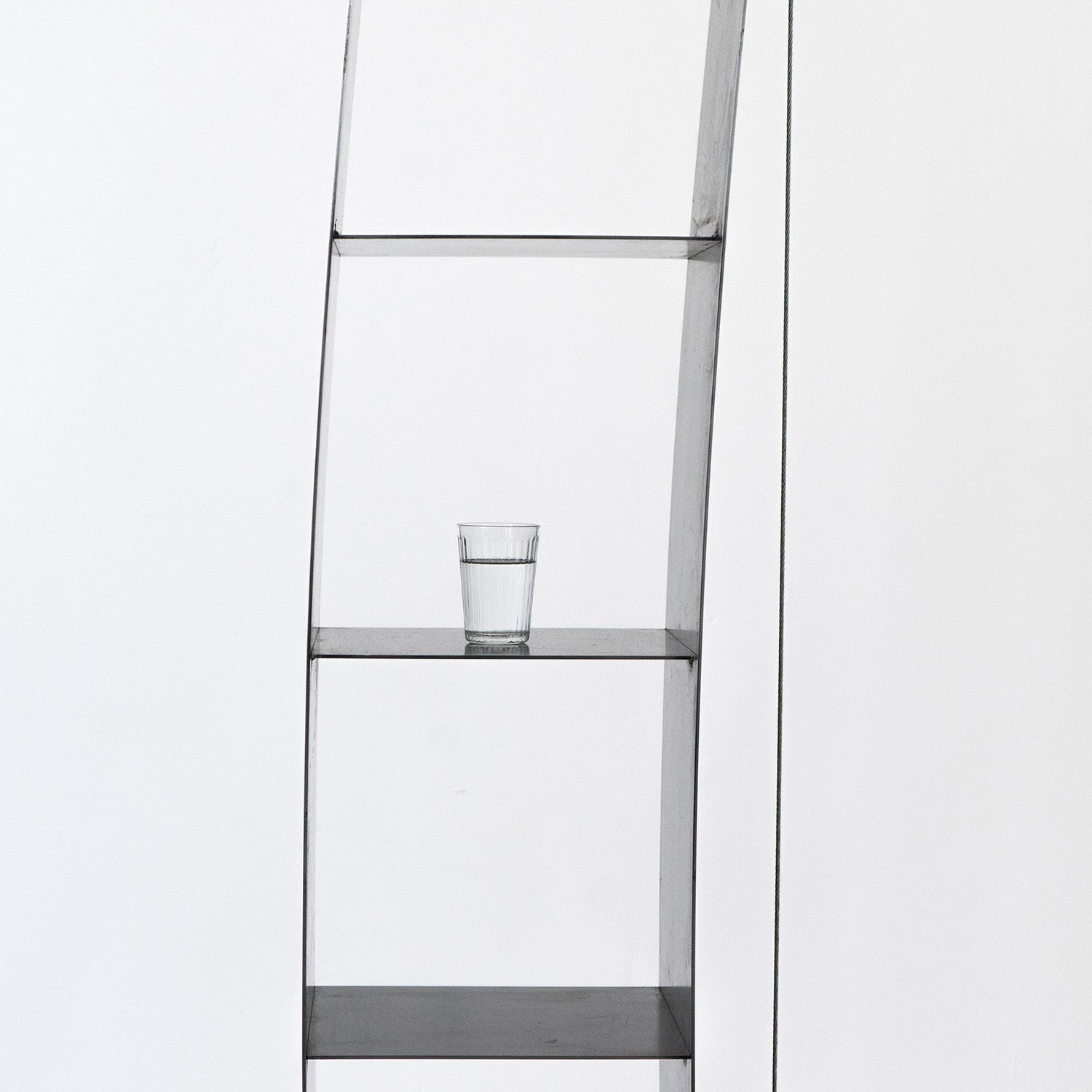 A modern Moormann Gespanntes Regal, with its minimalist design and sleek steel cables, is set against a white background. A partially filled glass of water rests on the second shelf, maintaining a simple and uncluttered composition.