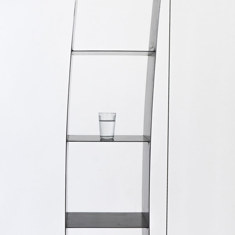 A modern Moormann Gespanntes Regal, with its minimalist design and sleek steel cables, is set against a white background. A partially filled glass of water rests on the second shelf, maintaining a simple and uncluttered composition.