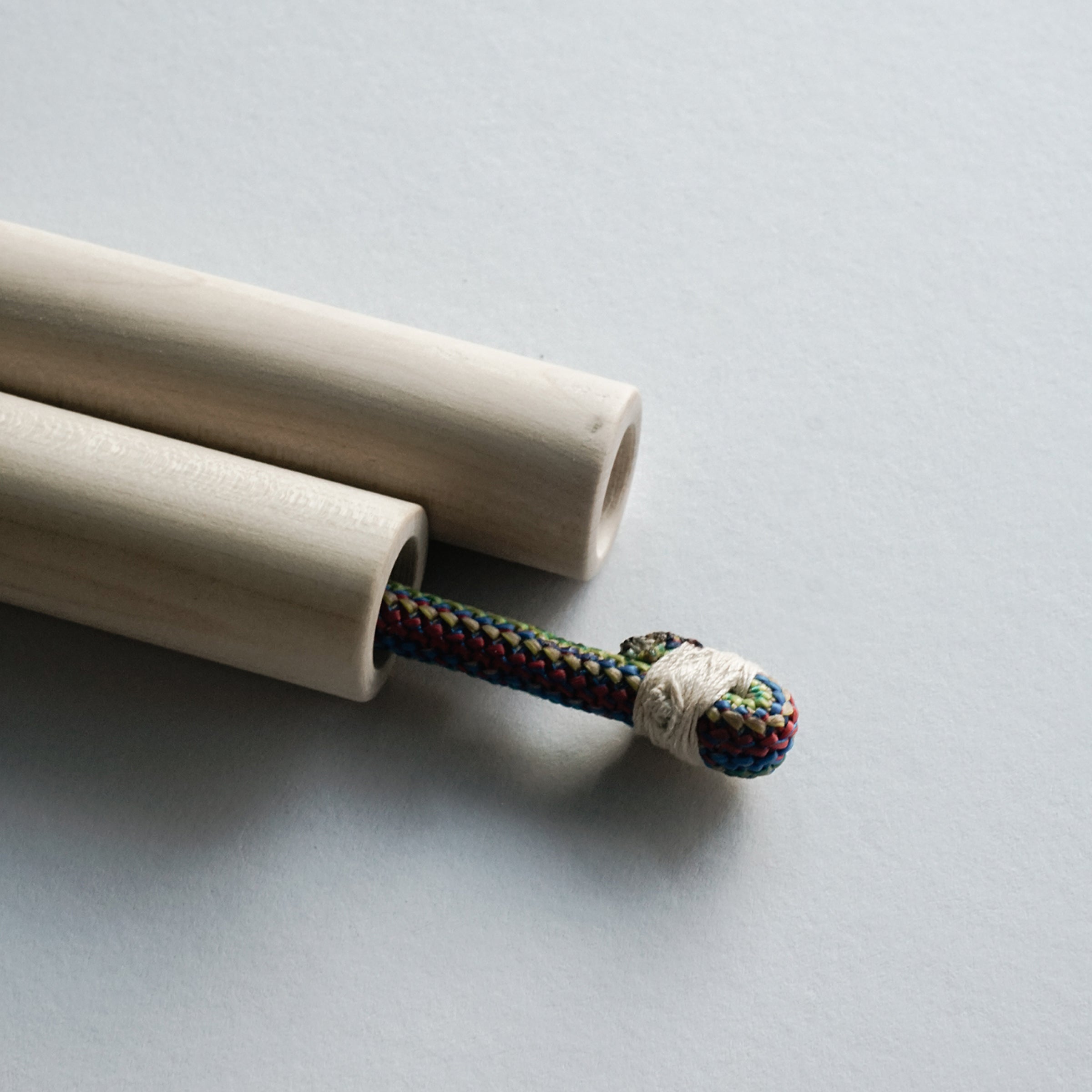 Two light wooden dowels parallel a colorful braided cord, wrapped with white string, crafted from ash and rope. This cheerful setup resembles the GoodGoods skipping rope, Spring, perfect for kids.