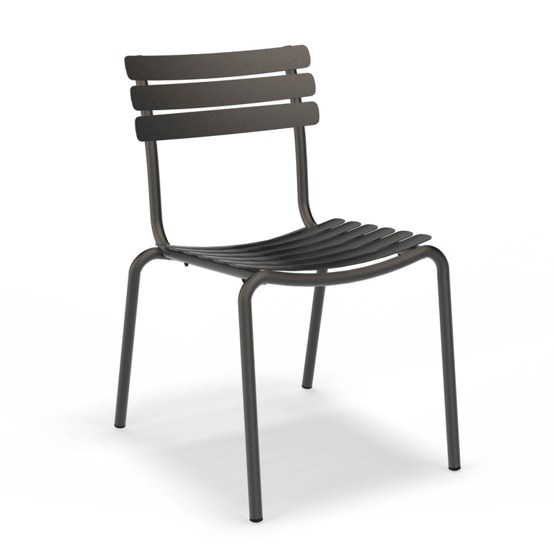 The Alua Dining Chair by Houe is a stackable, dark grey aluminum chair with a minimalist style. It features a slatted backrest and seat for versatile seating, while its four straight legs and slightly curved seat enhance comfort.