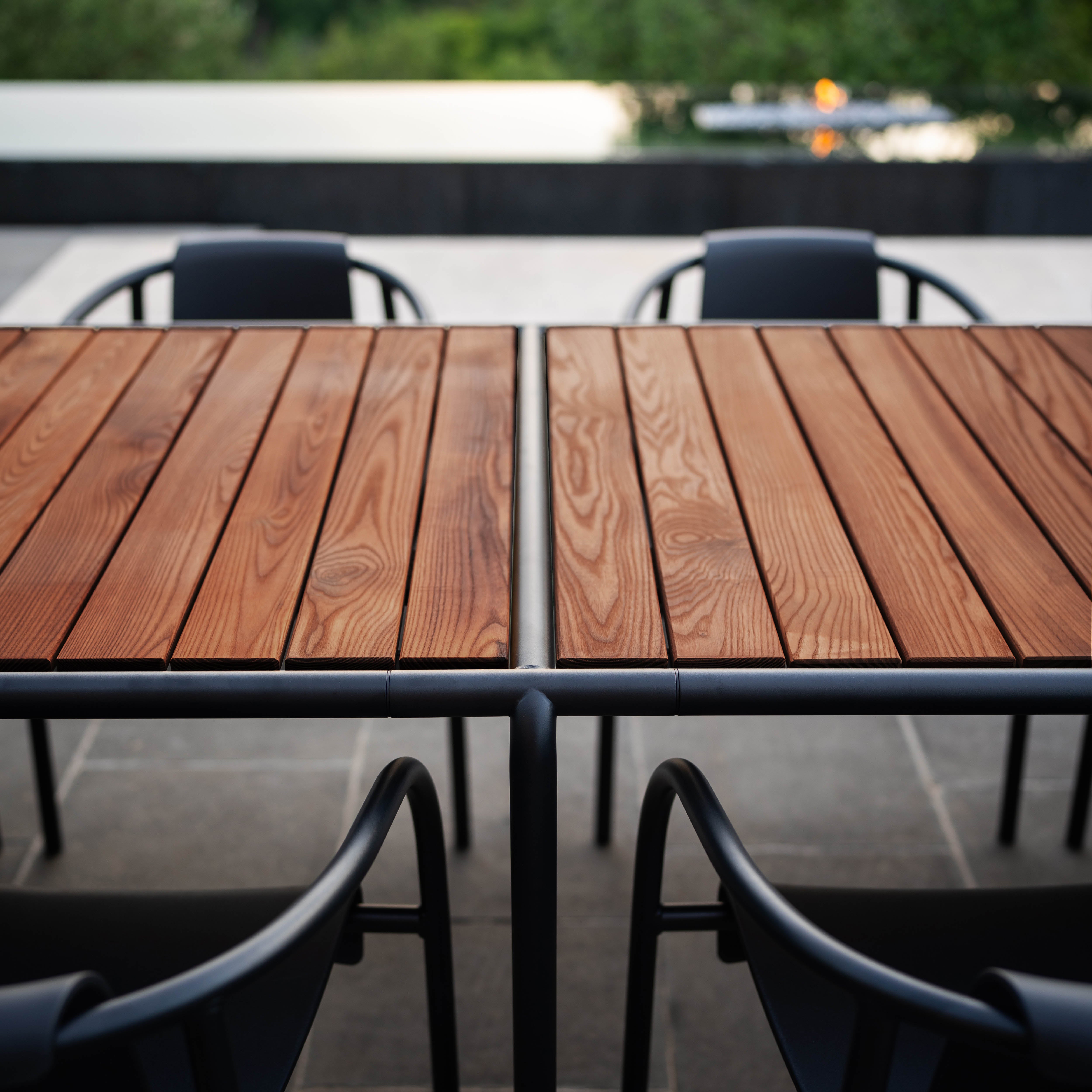 Modern outdoor dining setting with the AVANTI Table by Houe and black metal chairs on a stone-tiled patio. The sleek wooden table highlights its modular design and visible wood grain, while blurred greenery in the background complements the distant fire pit.