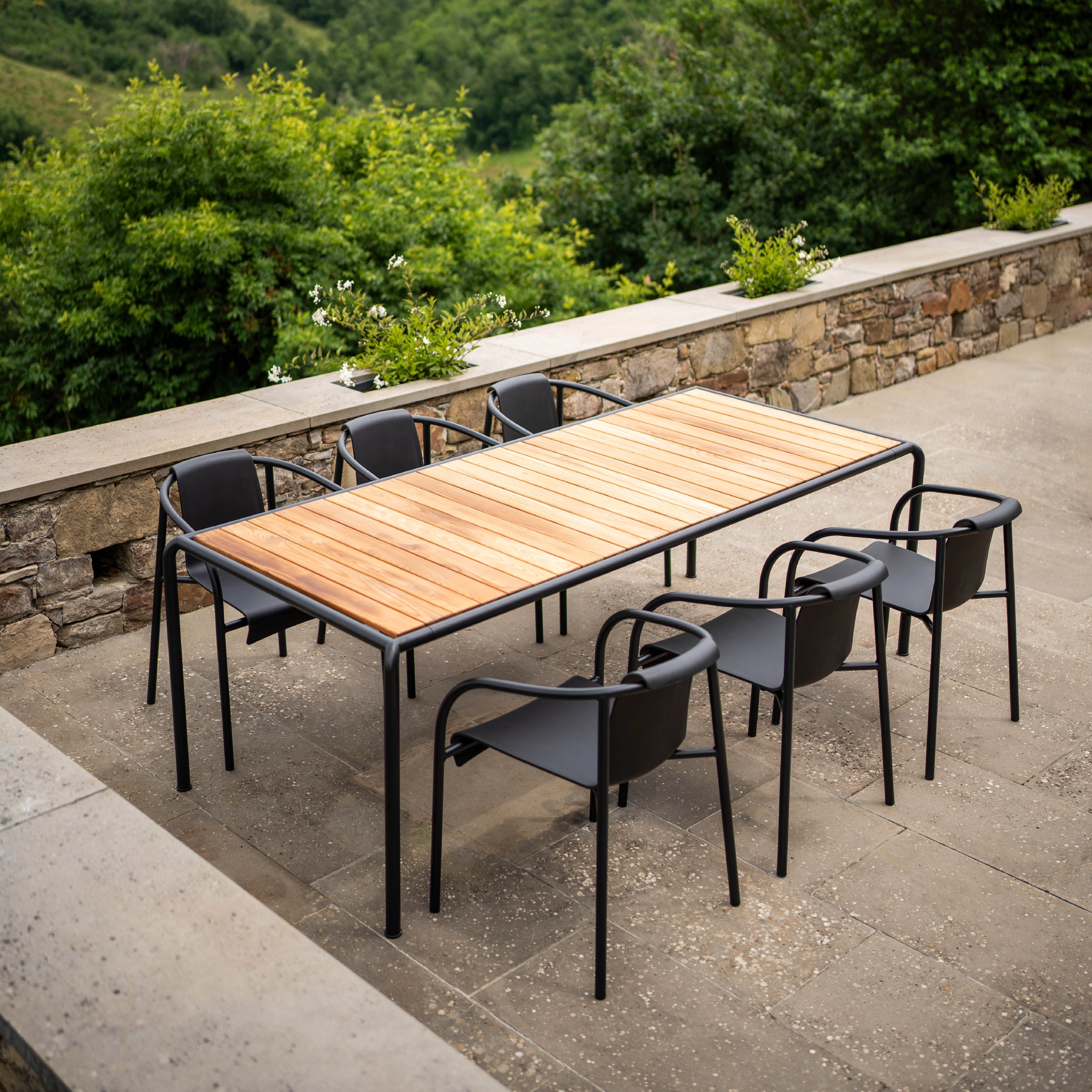 The outdoor dining area showcases a Houe Avanti Table paired with six black chairs on a stone patio. Lush foliage and hills create a serene atmosphere, enhanced by the tables modular design for versatile dining experiences.