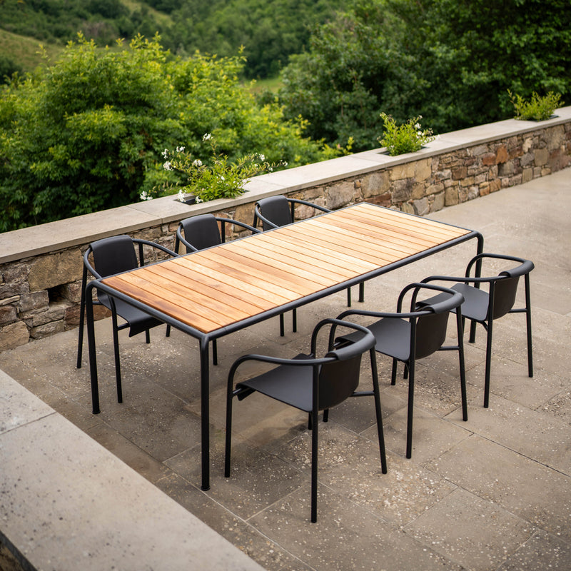 The outdoor dining area showcases a Houe Avanti Table paired with six black chairs on a stone patio. Lush foliage and hills create a serene atmosphere, enhanced by the tables modular design for versatile dining experiences.