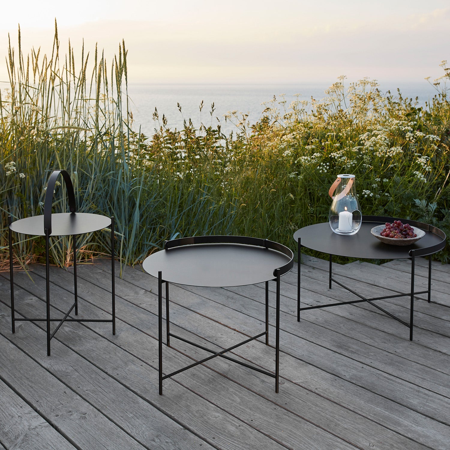 Three Edge Tray Tables by Houe, made of powder-coated black metal, sit on a wooden deck overlooking the sea. A glass pitcher with water is on one table, while another has a plate of grapes. Tall grasses and wildflowers border the deck under a clear sky with gentle, warm light.