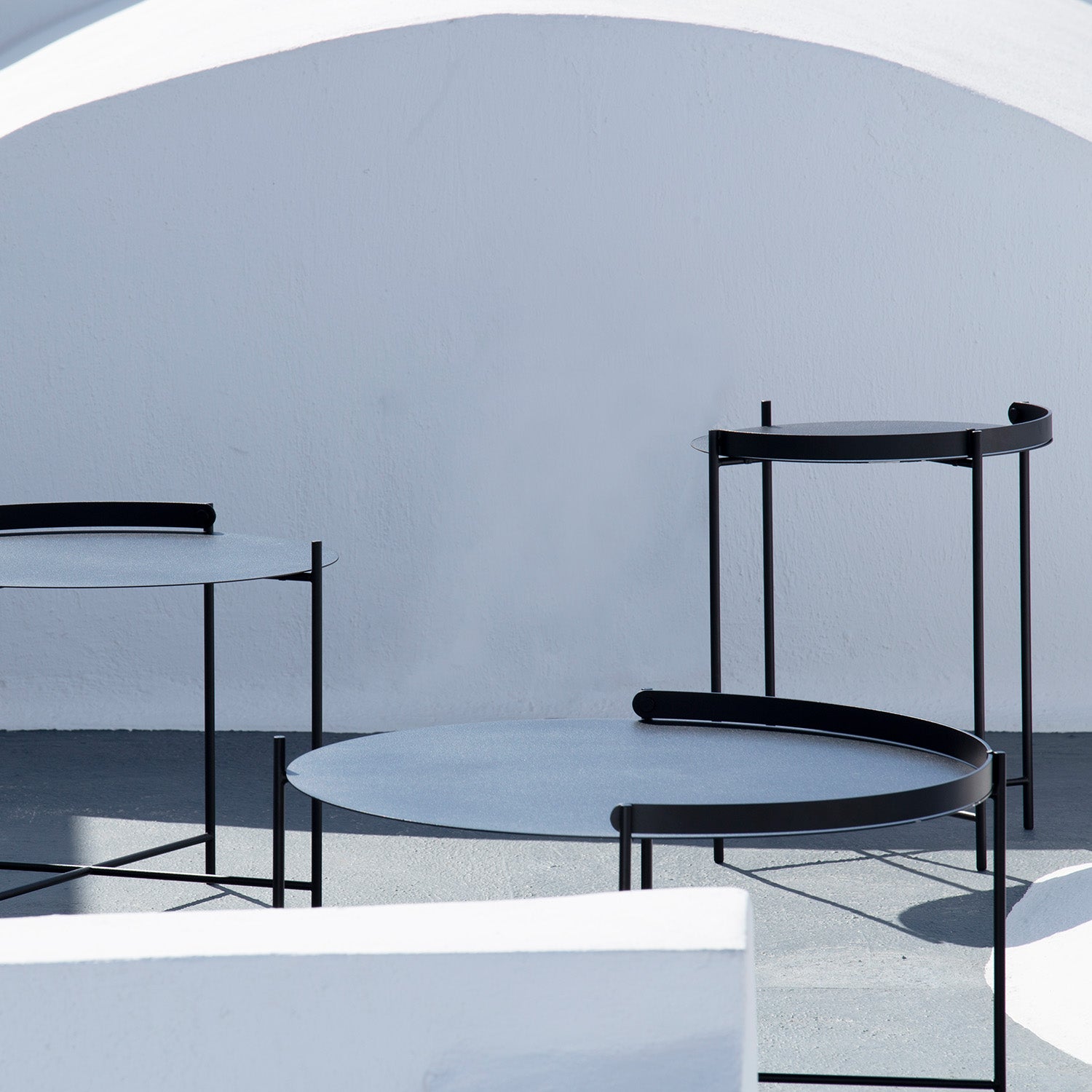 A minimalist scene includes three Edge Tray Tables from Houe, each with a modern round design and varying heights, set on a concrete floor. The tables feature black powder-coated steel frames and light-colored tabletops against a white curved architectural backdrop, casting soft shadows.