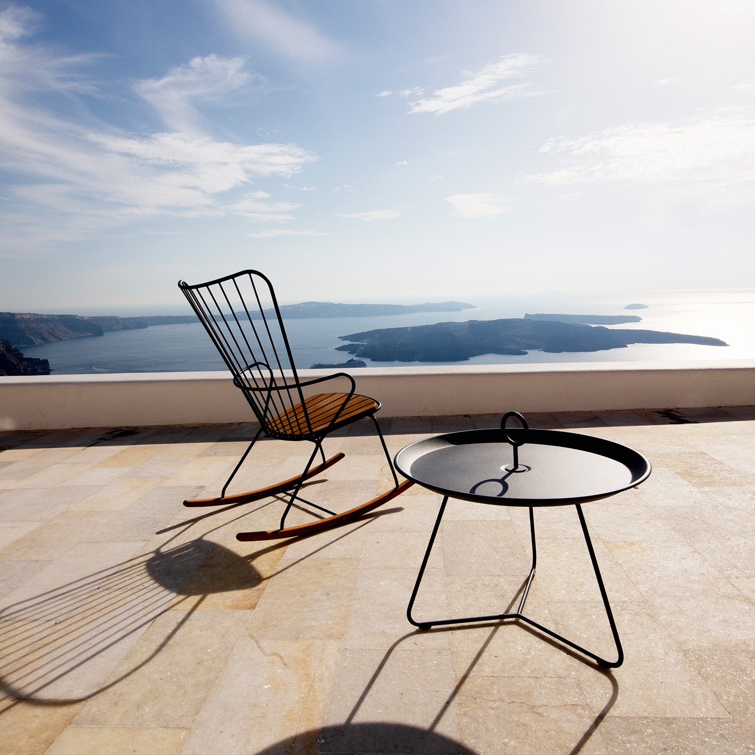 The Eyelet Tray Table by Houe and a metal rocking chair rest on a sunlit terrace overlooking a tranquil sea and distant islands under a partly cloudy sky. Shadows fall on the tiled floor, adding to the peaceful ambiance.