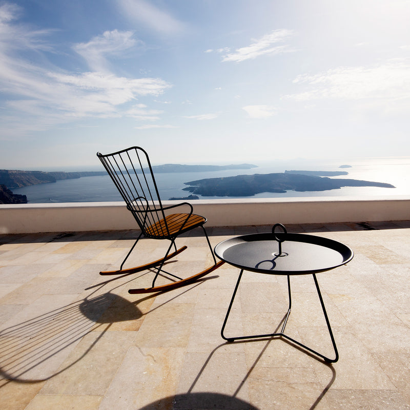The Eyelet Tray Table by Houe and a metal rocking chair rest on a sunlit terrace overlooking a tranquil sea and distant islands under a partly cloudy sky. Shadows fall on the tiled floor, adding to the peaceful ambiance.