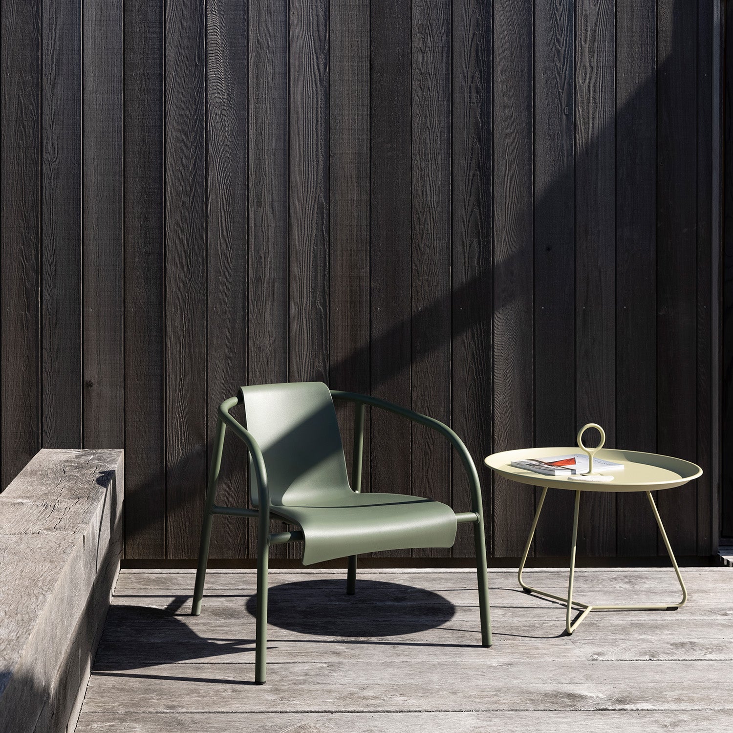 A modern outdoor scene features a green chair with metal arms beside Houes Eyelet Tray Table, a round, light yellow table made from powder-coated steel. A book and papers sit atop it, set against a dark wooden wall where sunlight casts intricate floor shadows.