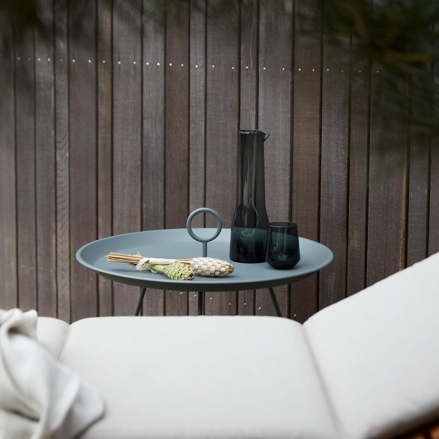 The Houe Eyelet Tray Table, a gray round side table made of powder-coated steel, elegantly supports a dark glass pitcher, two glasses, and a decorative flower with reeds. It complements a white lounge chair on the patio against a wooden fence, framed by green foliage.