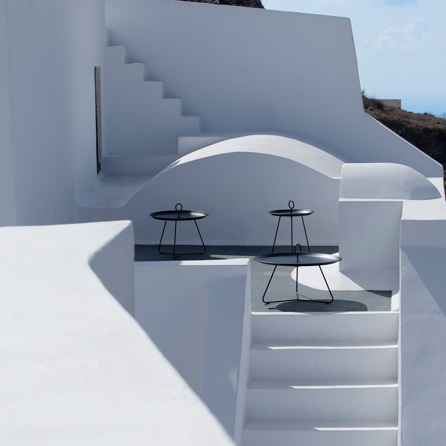 A minimalist outdoor space with white walls and stairs showcases two Houe Eyelet Tray Tables. These modern tables feature circular tops and tripod legs, crafted from powder-coated steel, enhancing the soft natural light for a serene, contemporary vibe.