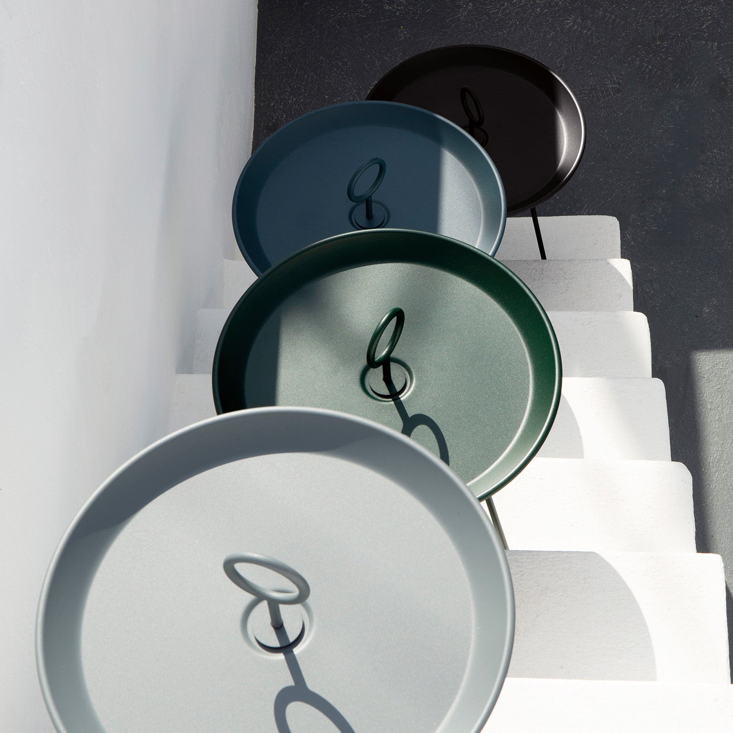 Houes Eyelet Tray Tables, crafted from powder-coated steel and featuring unique loop handles, are arranged diagonally on white stairs in shades of grey, blue, green, and black. These round tables create elegant shadows as they rest gracefully on each step.