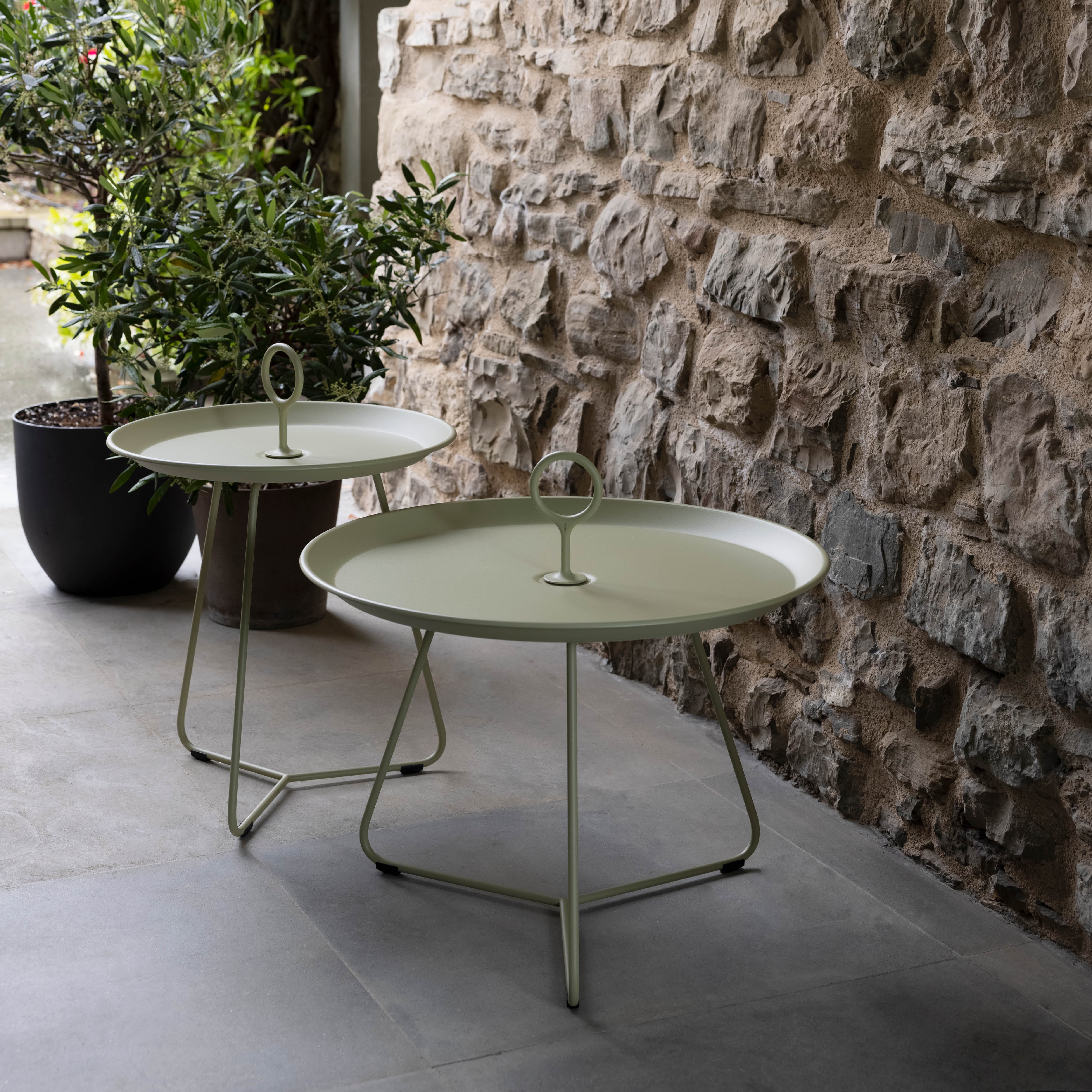 On a stone patio against a textured stone wall, two Houe Eyelet Tray Tables in light green powder-coated steel with handles stand. A potted plant with green leaves is partially visible in the background.