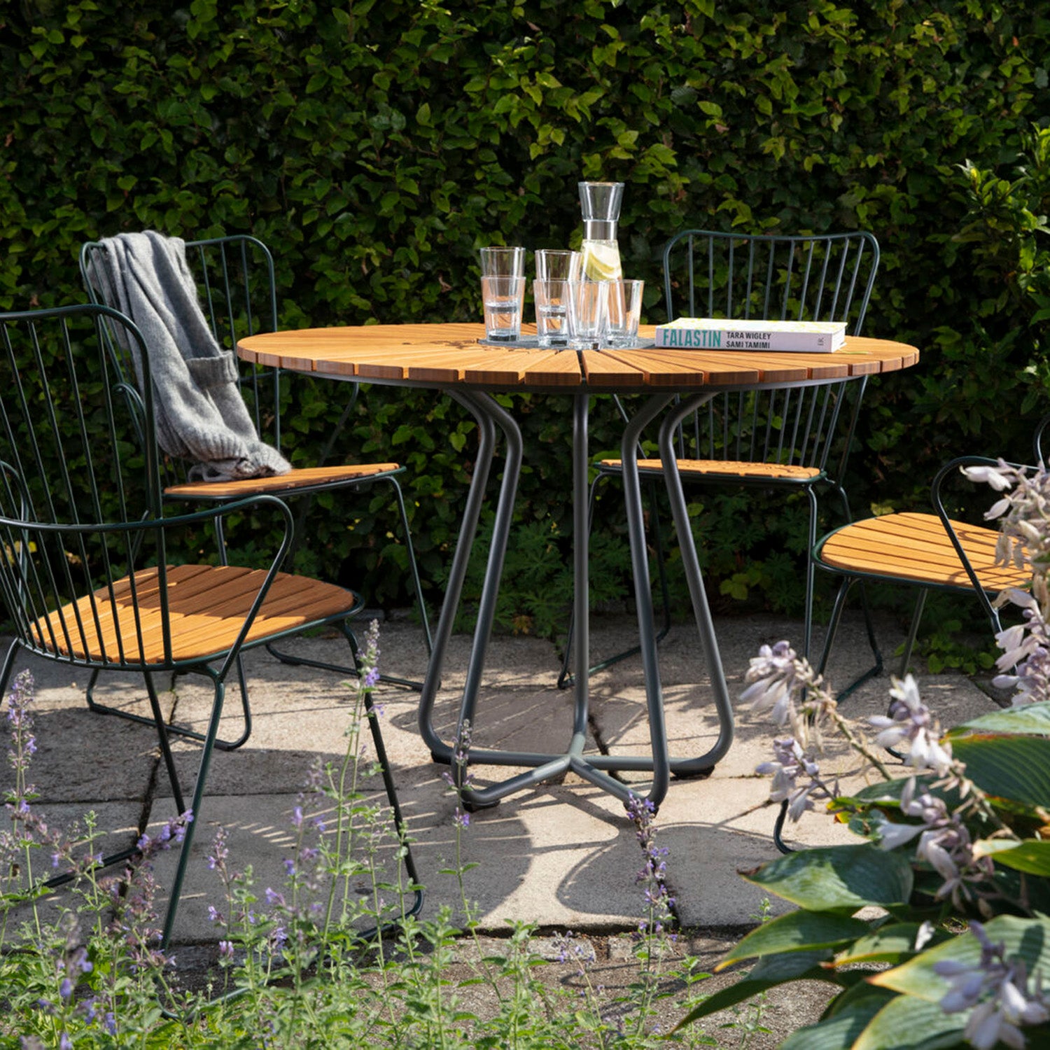 The Circle Table by Houe is set for outdoor dining with three matching chairs on stone tiles, featuring four glasses, a carafe, and a book. A gray blanket drapes one chair while green bushes and plants enhance the serene ambiance.