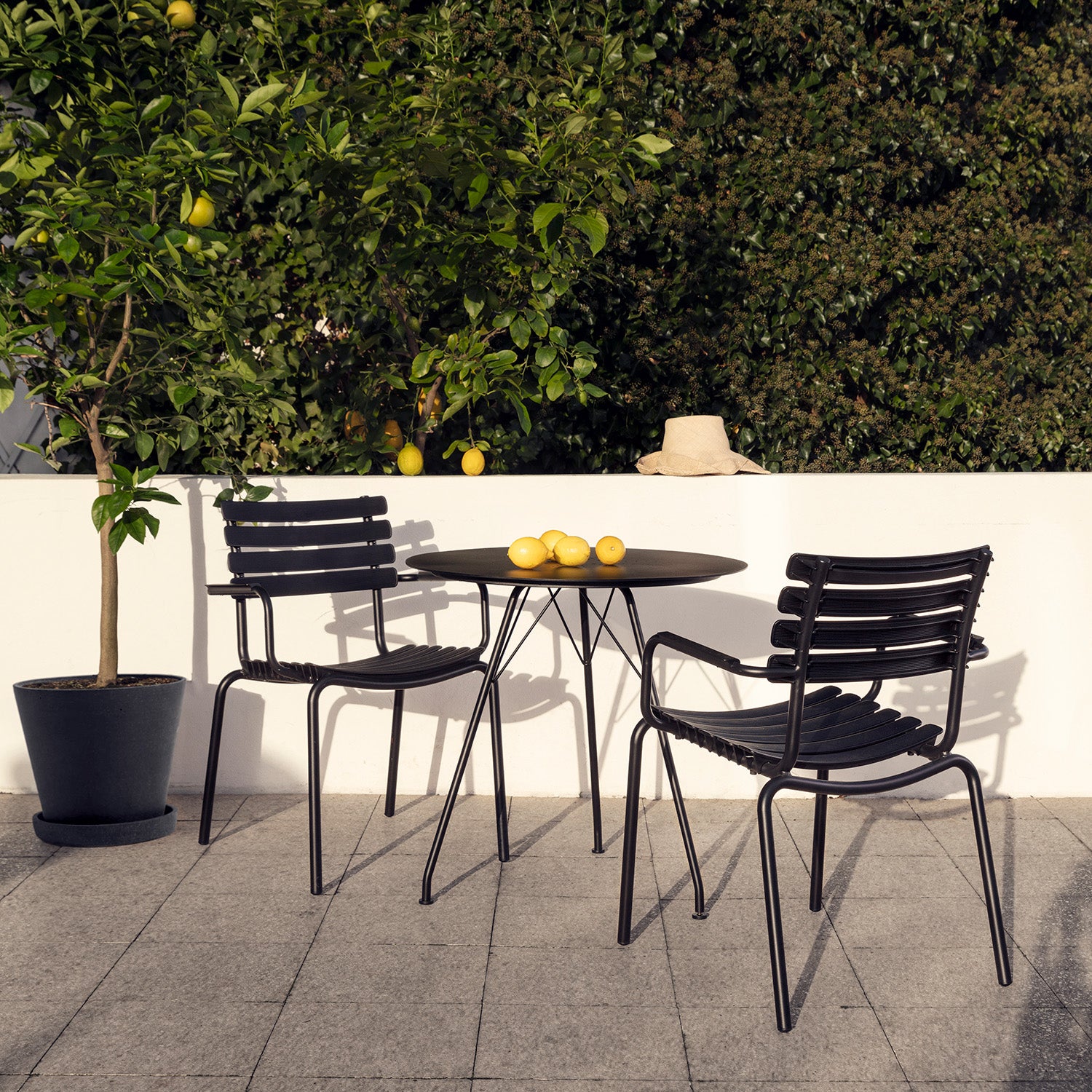 A cozy patio displays powder-coated steel dining chairs and a Houe Circum Table. Three lemons are placed on the table, beside a potted lemon tree full of fruit. A beige hat is on the white railing, contrasting with lush greenery in the background for a peaceful retreat.