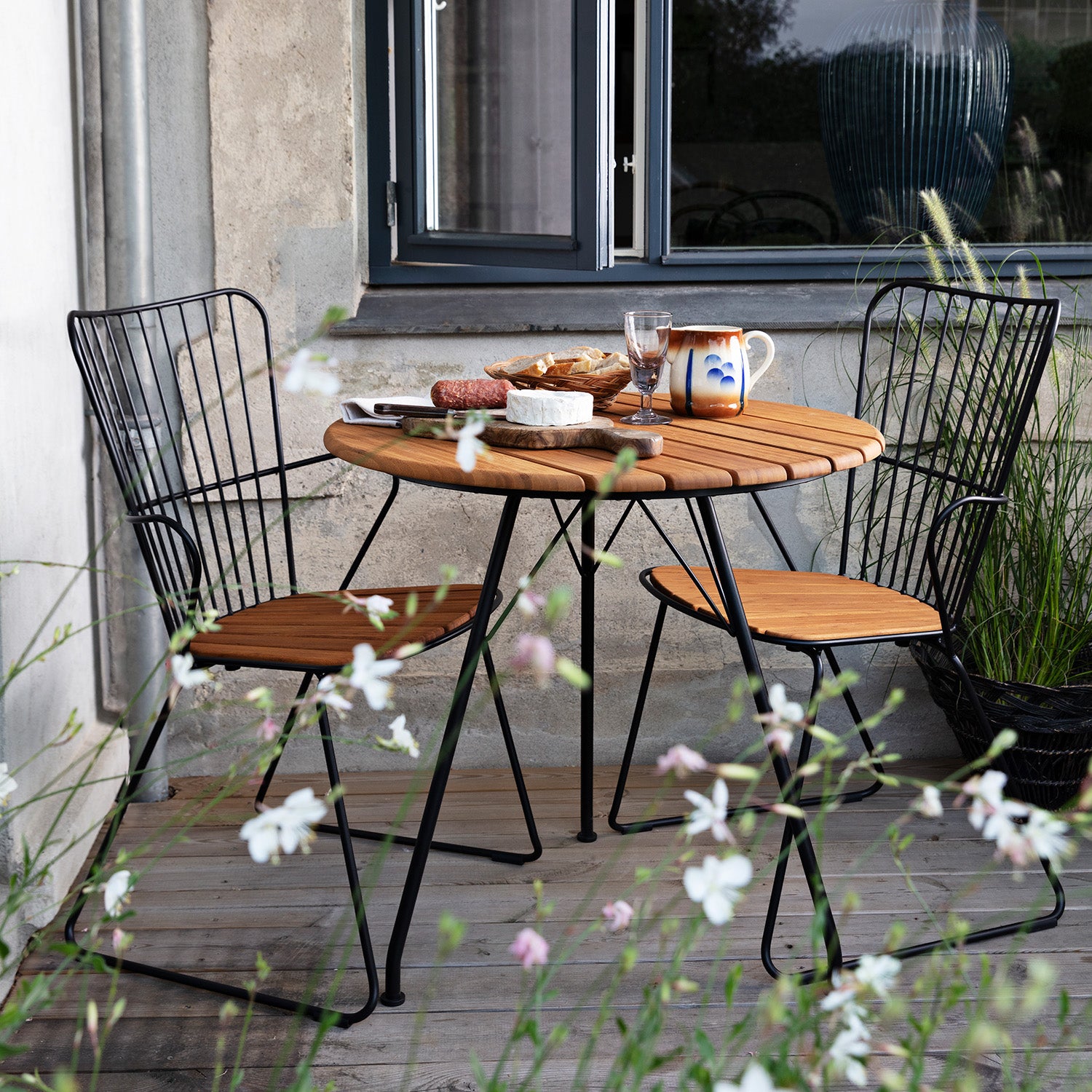 A charming outdoor dining scene showcases the Circum Table from Houe, paired with powder-coated steel HOUE dining chairs. The table is set for a meal, surrounded by tall white flowers against a gray building wall with a large window.