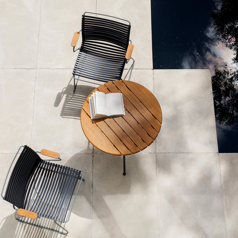 A round Circum Table with an open book is on a light-tiled patio by a sky-reflecting pool. Two Houe dining chairs with wooden armrests enhance the elegant and serene outdoor dining setting.