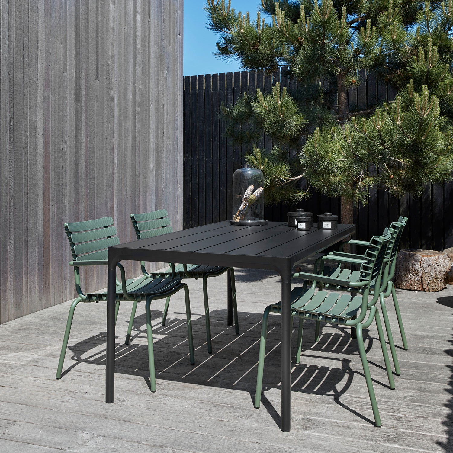 An outdoor patio features Houes Four Table in powder-coated aluminum, surrounded by six green chairs on a wooden deck. The area is bordered by bamboo-crafted fencing and a small tree. A glass cloche with an item inside and two small black containers are placed on the table.