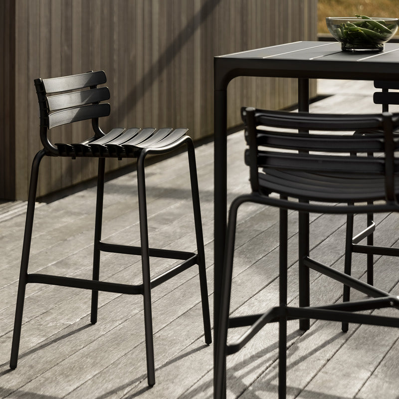 The outdoor dining area boasts Houes sleek, modern black metal furniture crafted from powder-coated aluminum. A high chair with horizontal slats pairs with the matching Four Bar Table on the wooden deck. Sunlit shadows play softly as a bowl of green beans adorns the table.