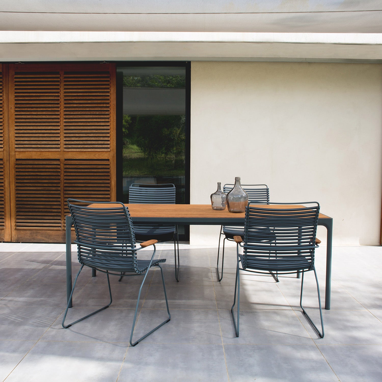 A minimalist patio features a Four Table by Houe, an elegant wooden dining table with four powder-coated aluminum chairs. Three decorative glass vases adorn the table, set against wooden shutters and a glass door.