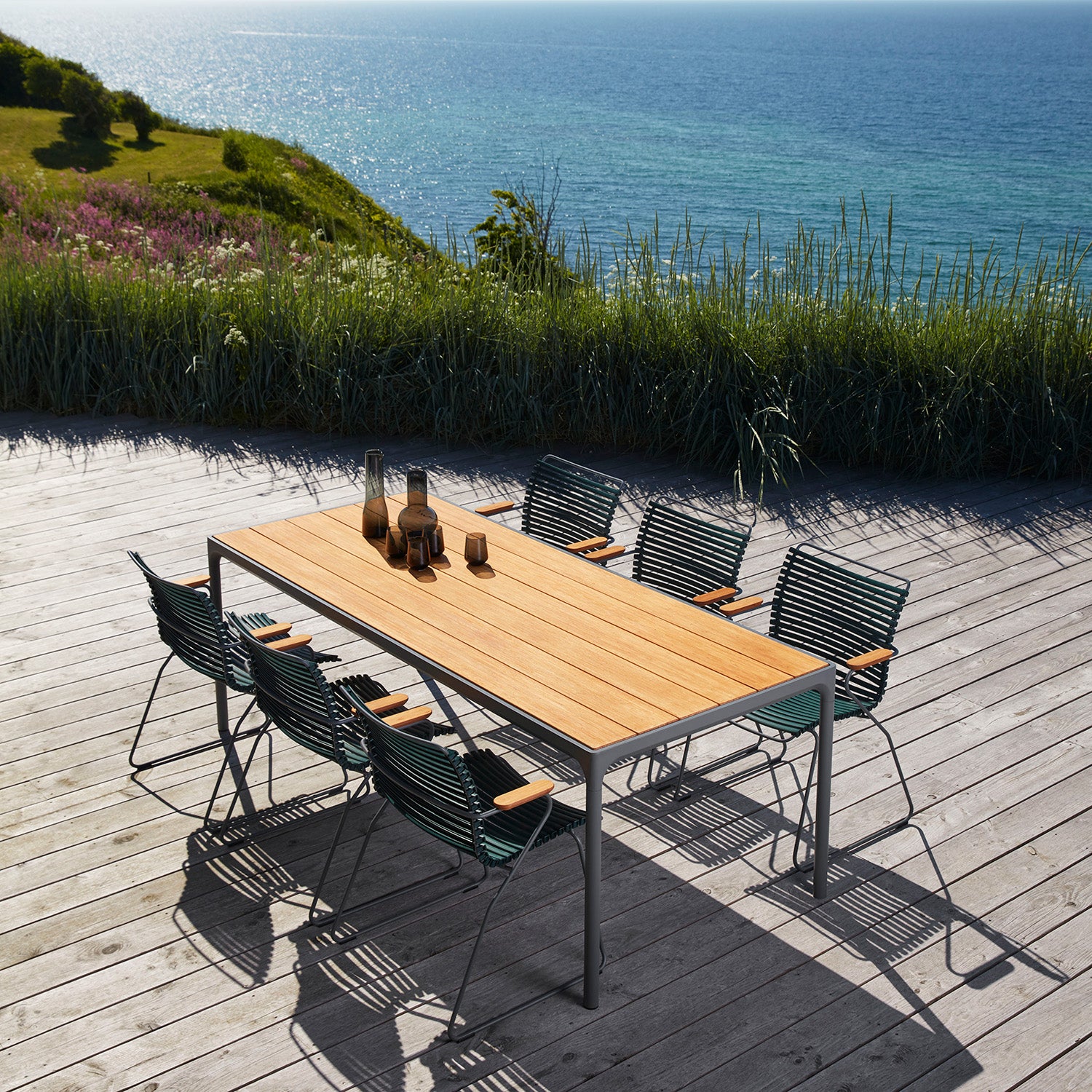 The Houe Four Table, a stunning wooden outdoor dining piece, is elegantly paired with eight chairs on the oceanfront deck. Three glass bottles and glasses enhance the setting, framed by lush grass and a clear blue sky, blending natural beauty with comfort effortlessly.