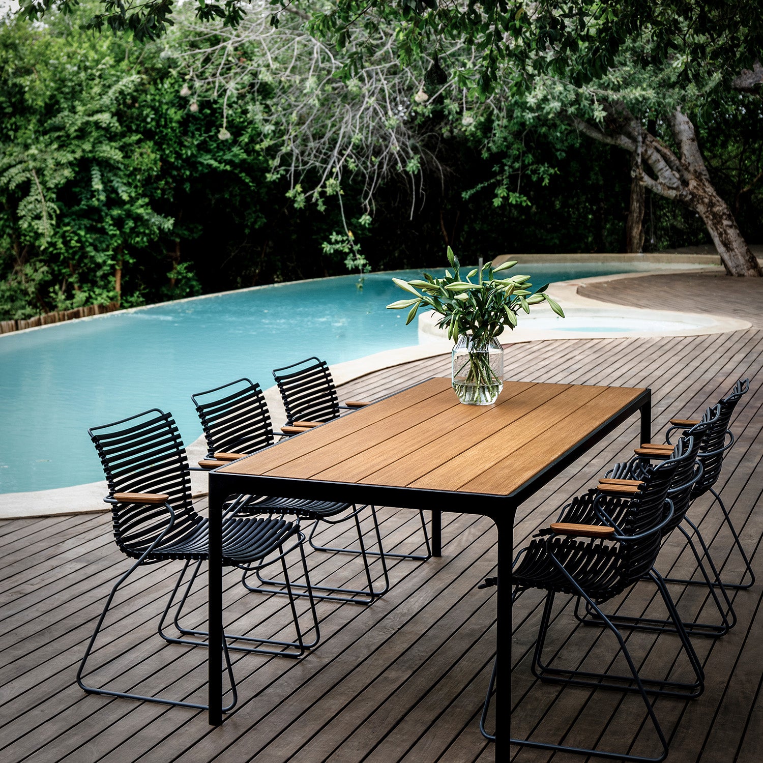 The outdoor dining setup by a curvy pool features Houes Four Table and six sleek black powder-coated aluminum chairs with wooden armrests. Set on a wooden deck, adorned with a vase of greenery, and surrounded by lush trees, it creates a serene and inviting atmosphere.