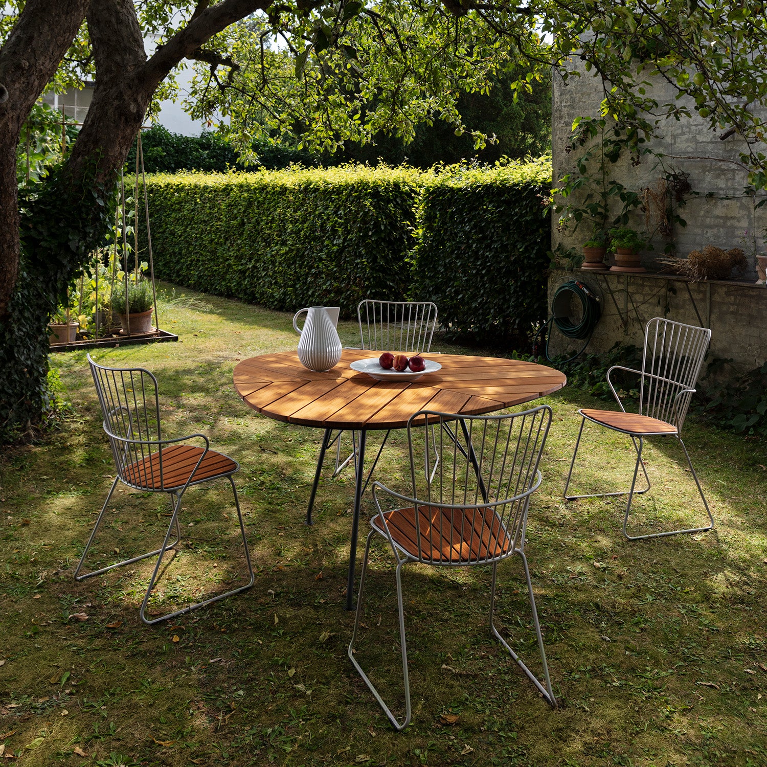 The Leaf Table by Houe, topped with a white pitcher and three apples, sits in a lush garden. Surrounded by four chairs with powder coated steel frames and wooden seats, its shaded by a tree and set against green hedges and a garden wall—perfect for serene outdoor dining.