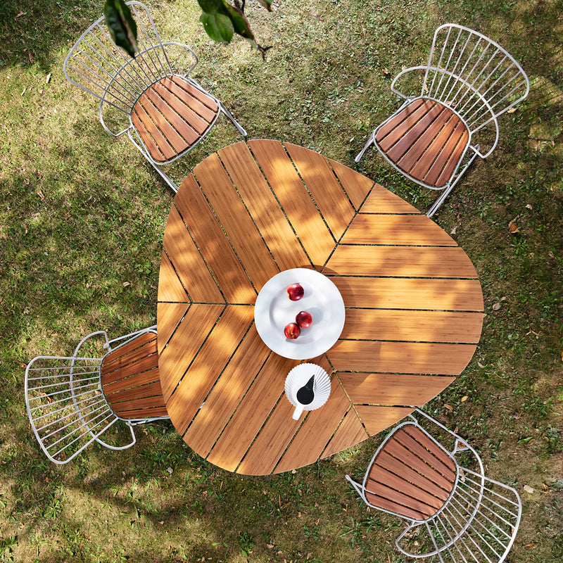 Aerial view of Houes Leaf Table, wooden and triangular with rounded edges, surrounded by four powder-coated steel chairs. On the table is a white plate with three red apples and a small black dish, set on grass—a perfect outdoor dining setup.