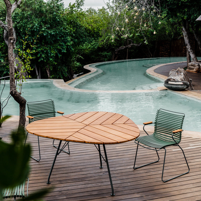 A tranquil outdoor setting showcases a kidney-shaped pool amidst lush greenery and bamboo. In the foreground, Houes Leaf Table with three powder-coated steel chairs sits on a wooden deck, enhancing the peaceful ambiance.