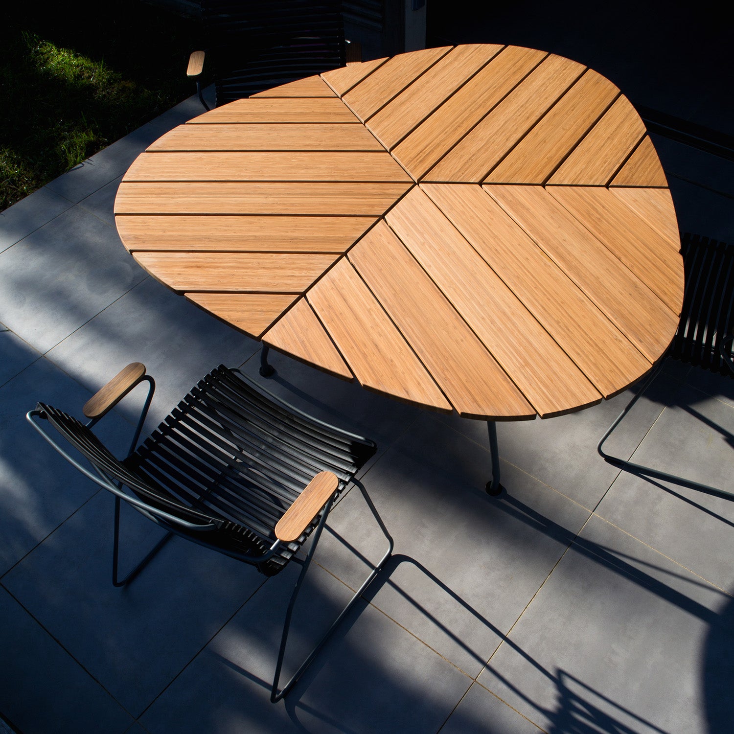 The Houe Leaf Table, a modern wooden outdoor dining table with a slatted design, stands prominently on the tiled patio. Its surrounded by minimalist black metal chairs with wooden armrests. Crafted from powder-coated steel, the furniture casts geometric shadows in the suns warm glow.