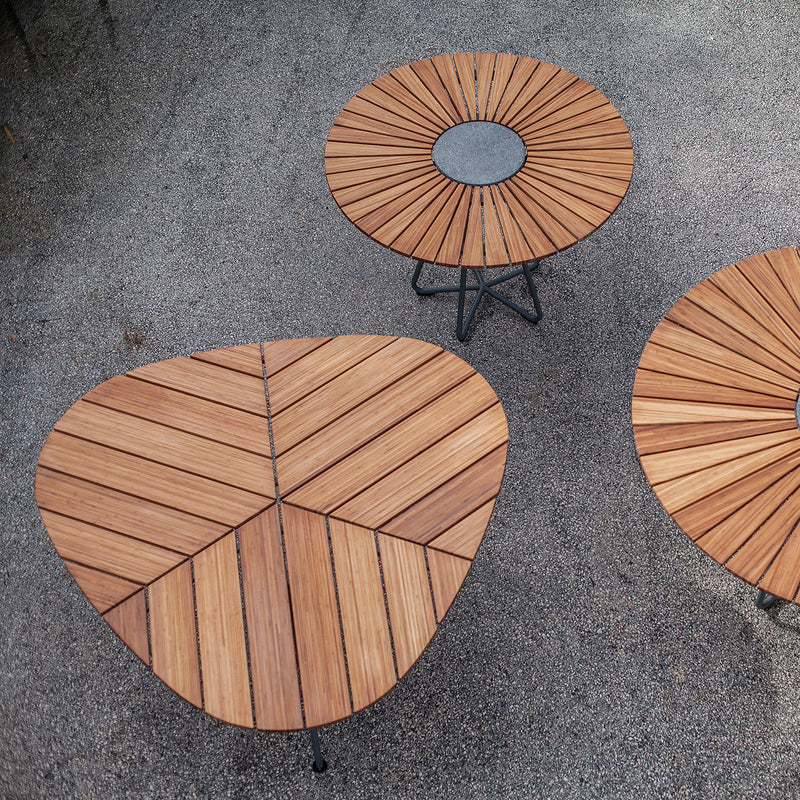 Three Leaf Tables by Houe sit on a gray pebble surface. The nearest bamboo table is triangular with chevron patterns, while the two round tables have powder-coated steel bases and central circles with radiating lines.