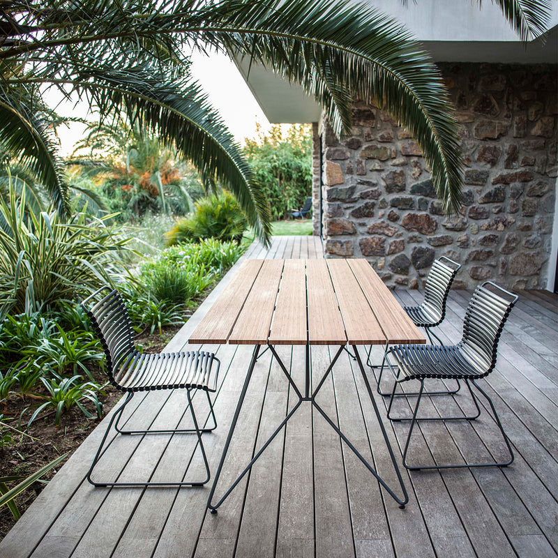 On a wooden deck, the Houe Sketch Table with four powder-coated steel chairs is surrounded by tropical plants next to a stone building. Palm fronds arch over the area, creating a serene setting perfect for outdoor dining.