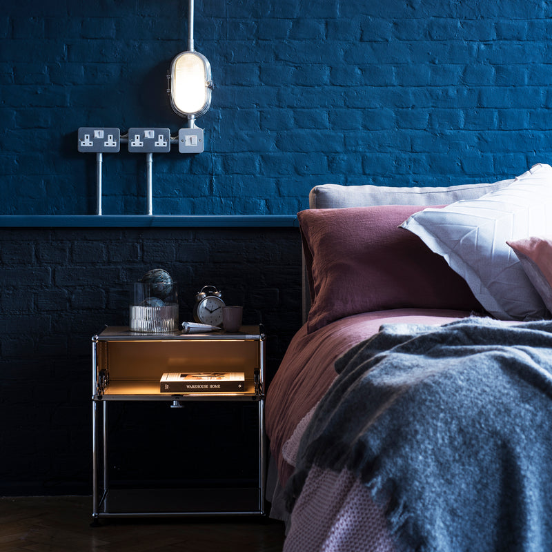 Haller Nightstand P2 in USM Beige with chrome frame, 6" drop-down lockable door next to a bed with blue wall background.