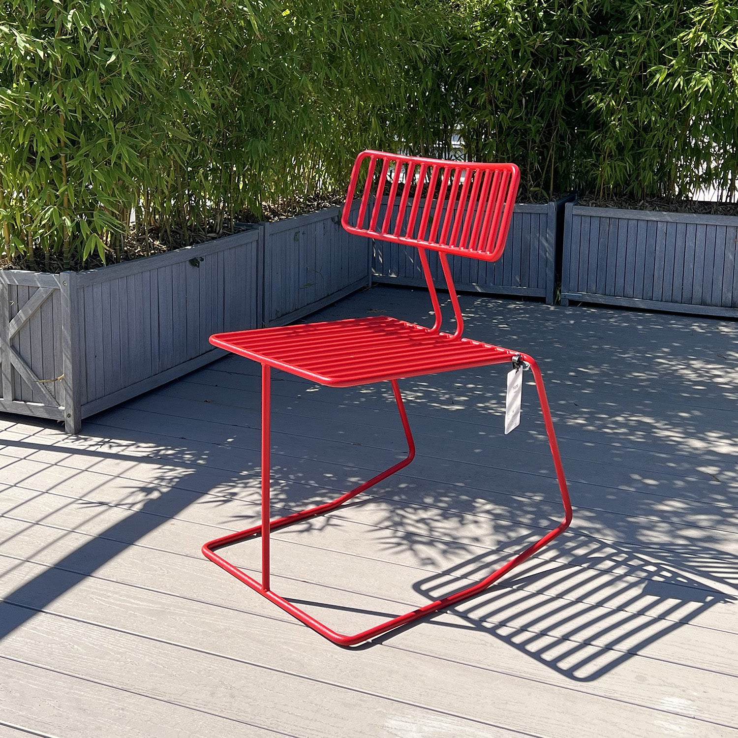 Egoes Hope Chair - SALE, boasts a modern red metal design that casts striped shadows on the gray deck. Nestled among tall green plants in rectangular planters, its stackable structure offers both comfort and convenience under bright sunlight.