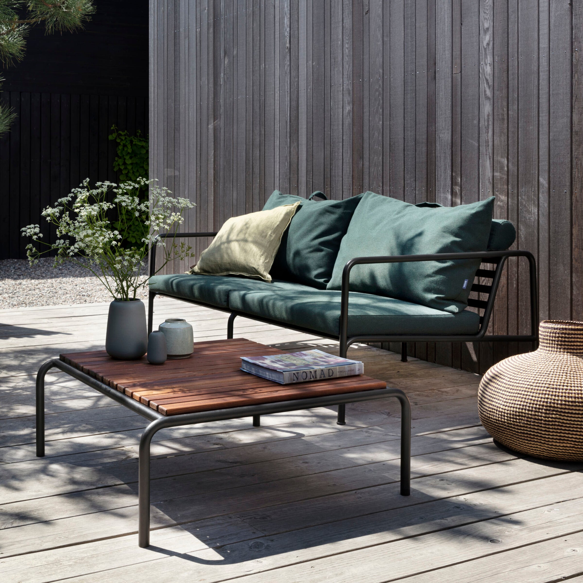 Outdoor patio with a weather-resistant metal-framed sofa featuring green cushions, an Avon Coffee Table by Houe, and decorative vases. A wicker basket sits on the right, creating a perfect lounge collection ambiance against the wooden fence on the deck.