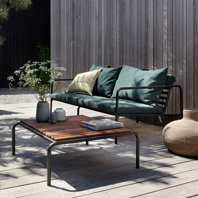 Outdoor patio with a weather-resistant metal-framed sofa featuring green cushions, an Avon Coffee Table by Houe, and decorative vases. A wicker basket sits on the right, creating a perfect lounge collection ambiance against the wooden fence on the deck.