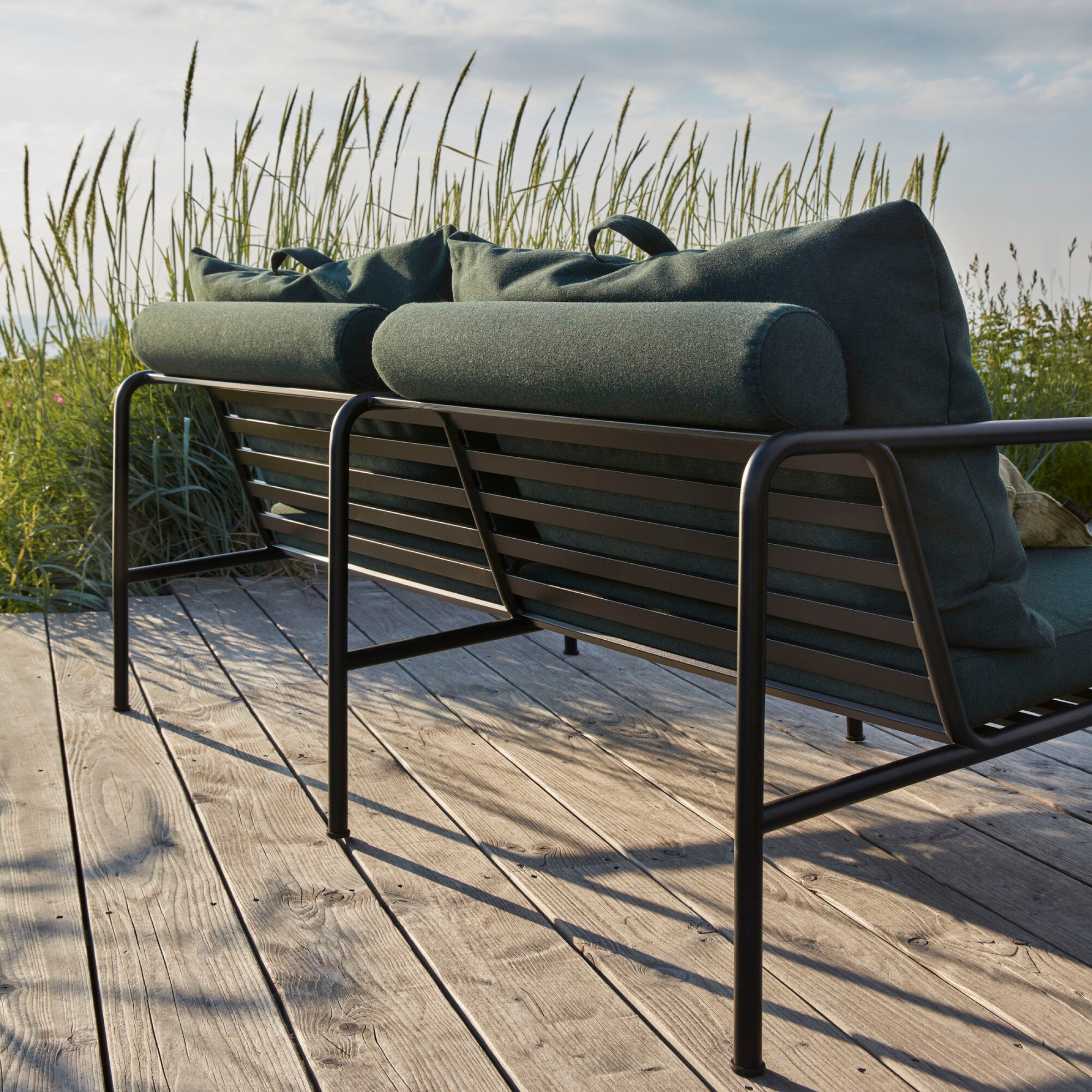 The Avon 2 Seater Sofa by Houe boasts dark, weather-resistant cushions on a wooden deck, overlooking a grassy area under a cloudy sky. Tall grass and wildflowers surround the setting, crafting a serene and natural atmosphere.