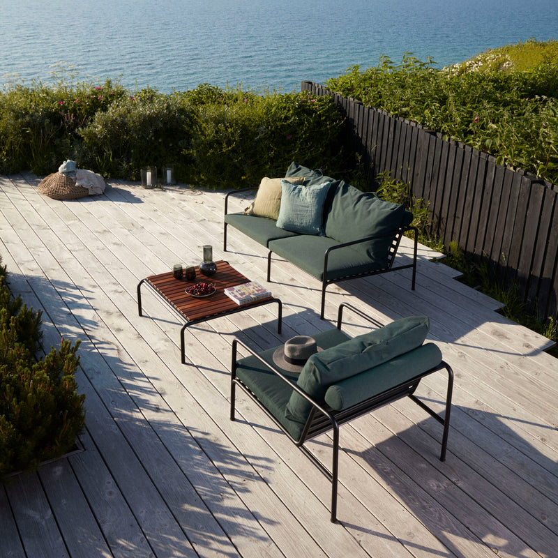 The patio, part of Houes AVON collection, features an Avon 2 Seater Sofa with weather-resistant green cushions, wooden coffee table, and chairs. It boasts a wooden deck overlooking serene waters, lush plants, and a black fence with a hat and two glasses on the lounge furniture.