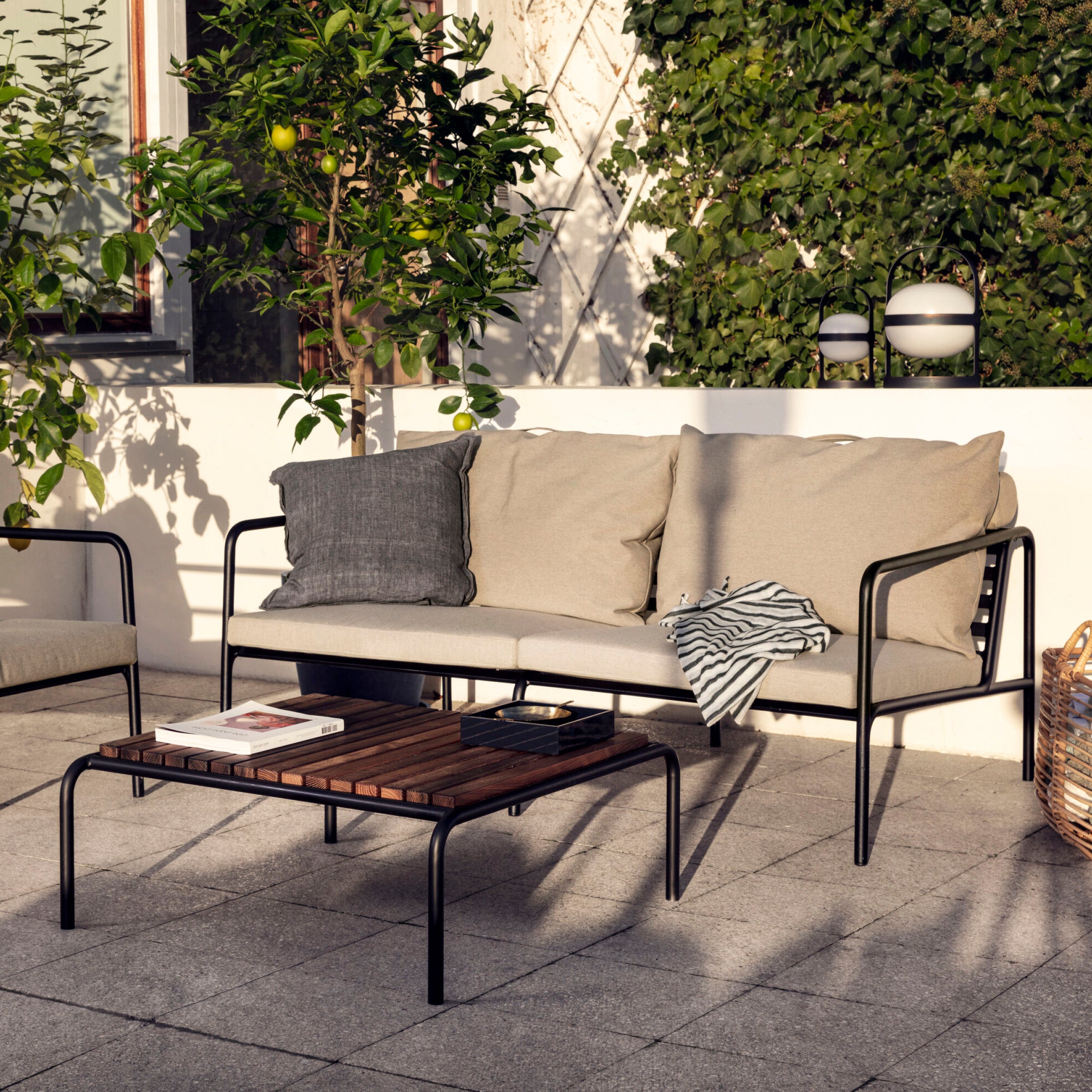 Outdoor patio featuring the Houe Avon 2 Seater Sofa in beige, with two throw pillows, a striped blanket, and a black cushion. A wooden coffee table displays two magazines. The weather-resistant setting is shaded by lush green plants.