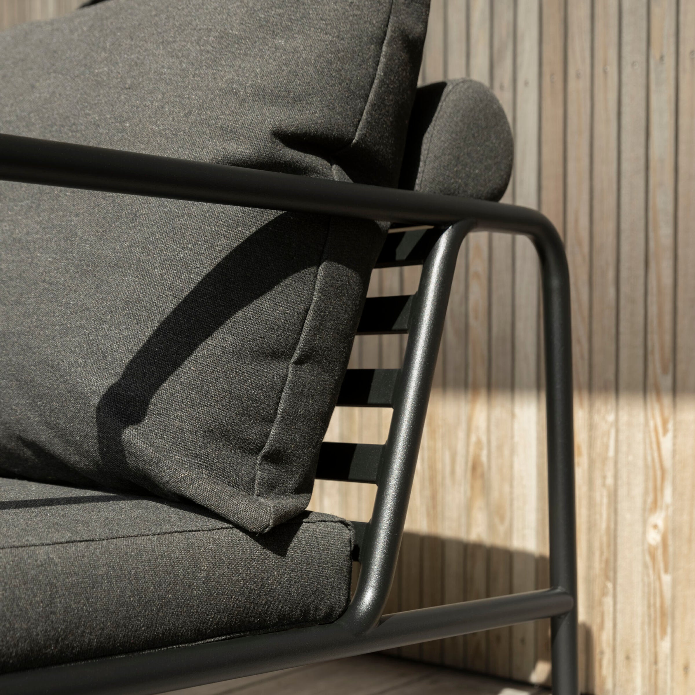 The AVON 2 Seater Sofa by Houe features dark gray cushions and a sleek black metal frame. Its weather-resistant and is shown against a wooden slat wall with sunlight casting gentle shadows on its surfaces.
