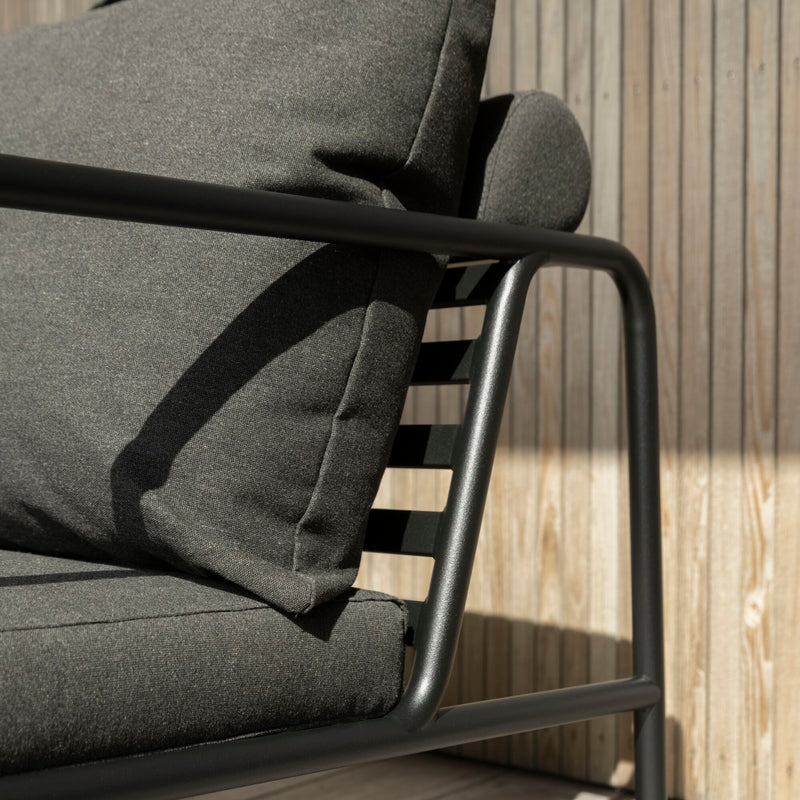 The AVON 2 Seater Sofa by Houe features dark gray cushions and a sleek black metal frame. Its weather-resistant and is shown against a wooden slat wall with sunlight casting gentle shadows on its surfaces.