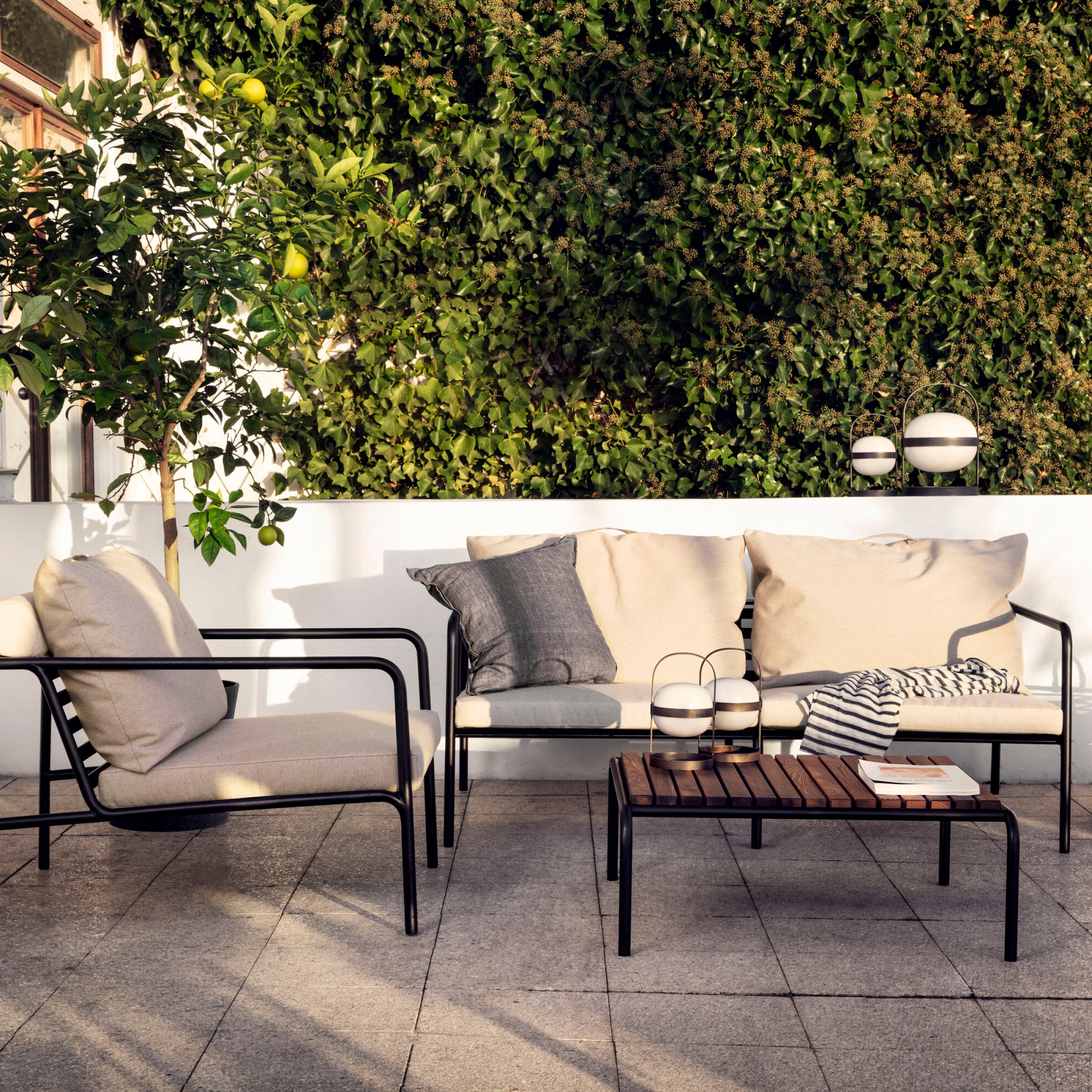 The patio showcases Houes weather-resistant Avon Lounge Chair with black metal and white cushions. A lemon tree stands left and green ivy forms a lush backdrop. A wooden coffee table is stylishly adorned with books and two lanterns.