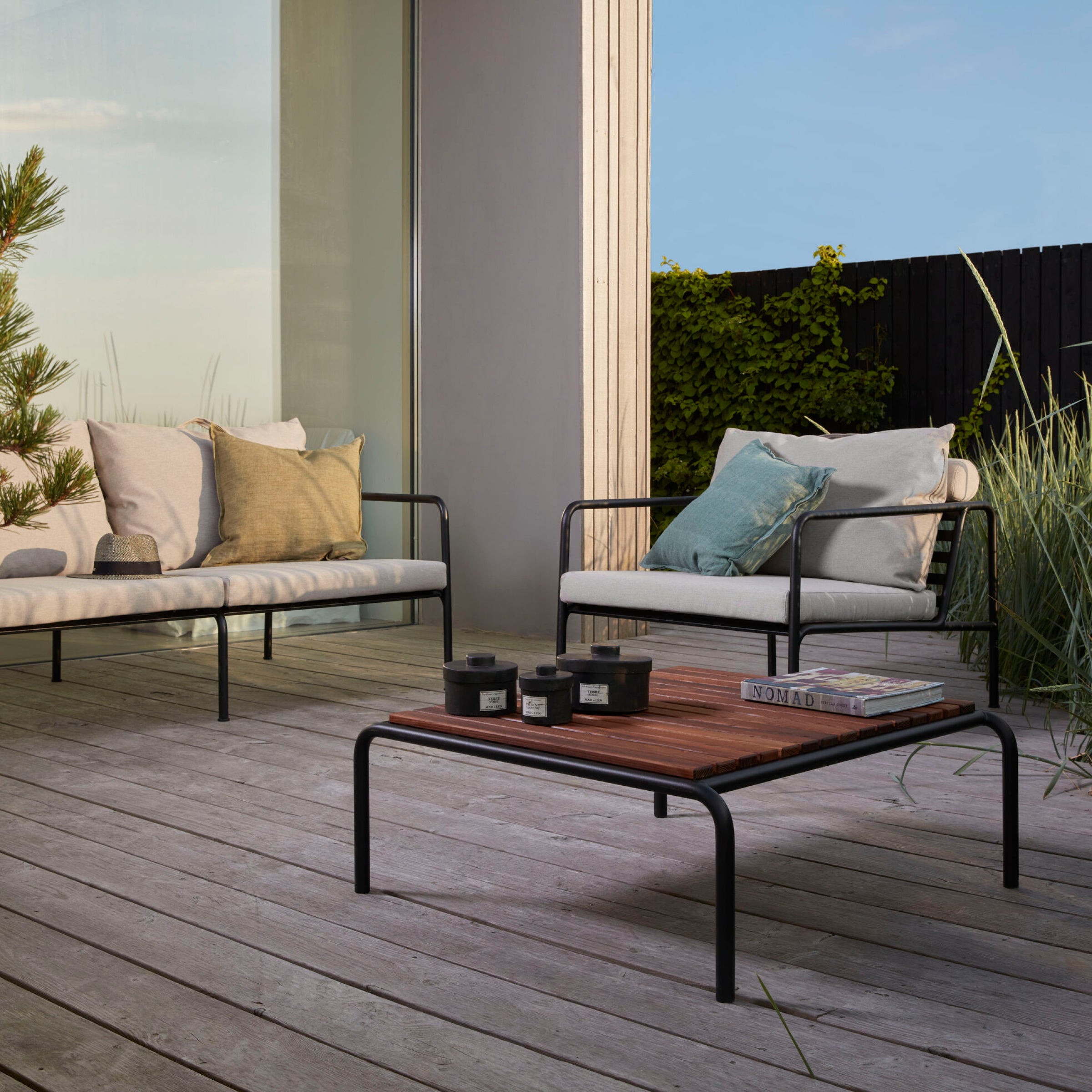 The outdoor patio showcases the AVON collection with two cushioned metal sofas, a Houe Avon Coffee Table, and decorative plants. Modern minimalist decor complements the wooden deck floor and large glass window, enhancing the serene, open vibe.