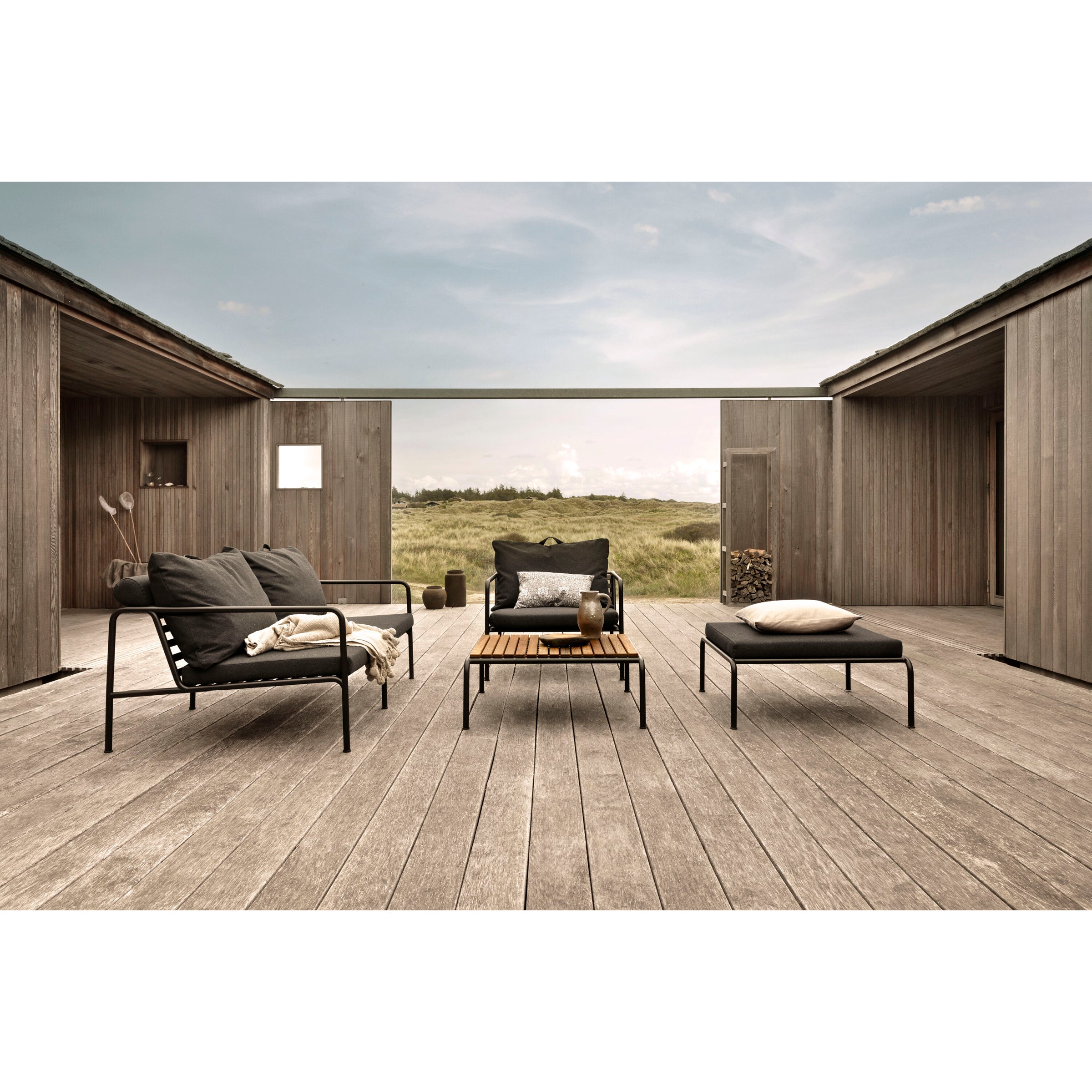 Explore the modern outdoor patio featuring two weather-resistant black chairs with cushions, a wooden coffee table, and the Avon Ottoman from Houe on a wooden deck. Wooden walls surround this space, offering views of a grassy field under a partly cloudy sky.