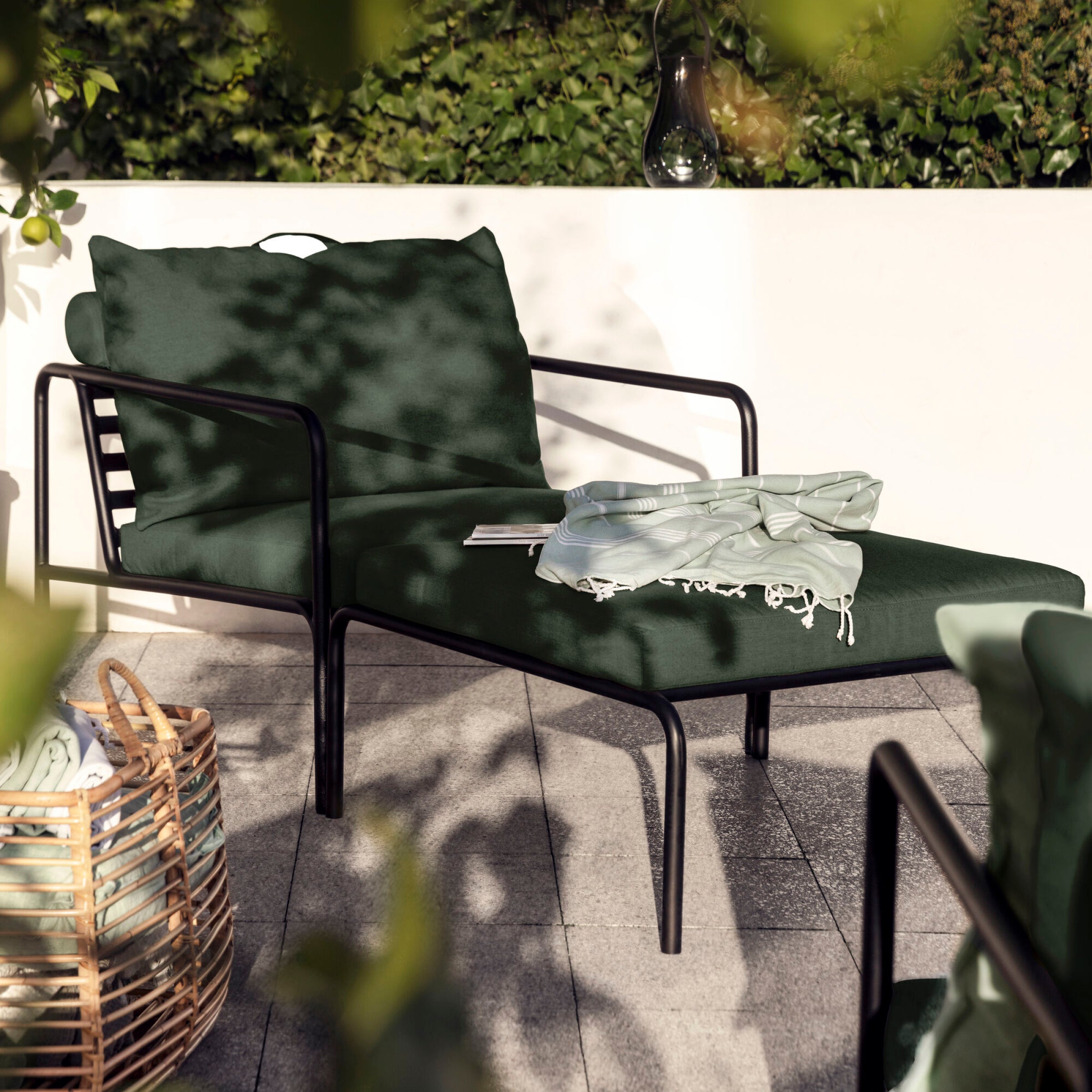 From the Houe AVON collection, this stylish outdoor ottoman features weather-resistant dark green cushions on a stone-tiled patio. A book and light scarf rest nearby, enhancing the tranquil setting with a wicker basket holding a blanket and lush plants.