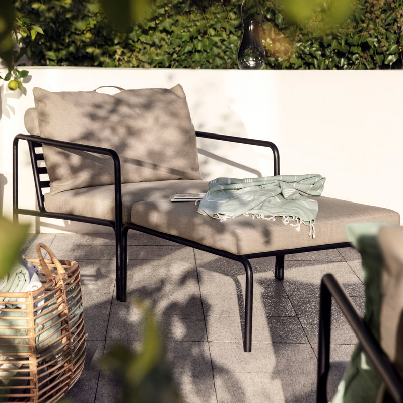 The Avon Lounge Chair by Houe, featuring UV-protected beige cushions, relaxes on a sunlit patio amidst vibrant greenery. A light blue scarf and a book rest on the chair while a wicker basket sits close by.