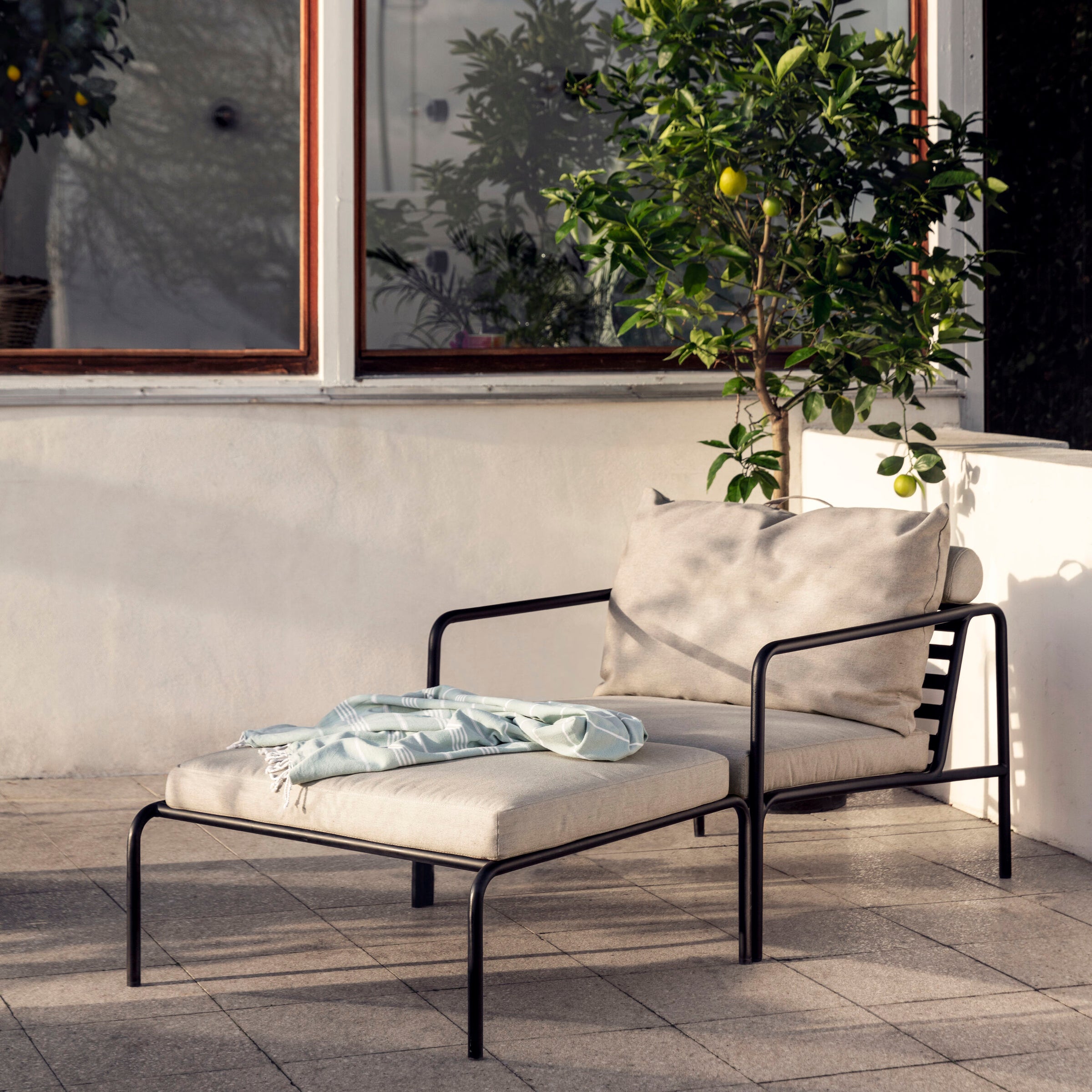 The Houe AVON collections outdoor lounge features a chic black metal-framed chair with beige cushions and an Avon Ottoman. Draped with a light blue blanket, it sits near a small lemon tree thriving against the light-colored walls with a window view.