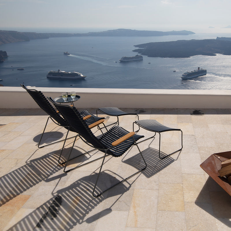 Two Houe Click Footrests, crafted with UV-protected plastic lamellas, flank a small round table with drinks on a stone terrace overlooking a serene ocean view dotted with cruise ships. The footrests cast elegant shadows on the terrace.