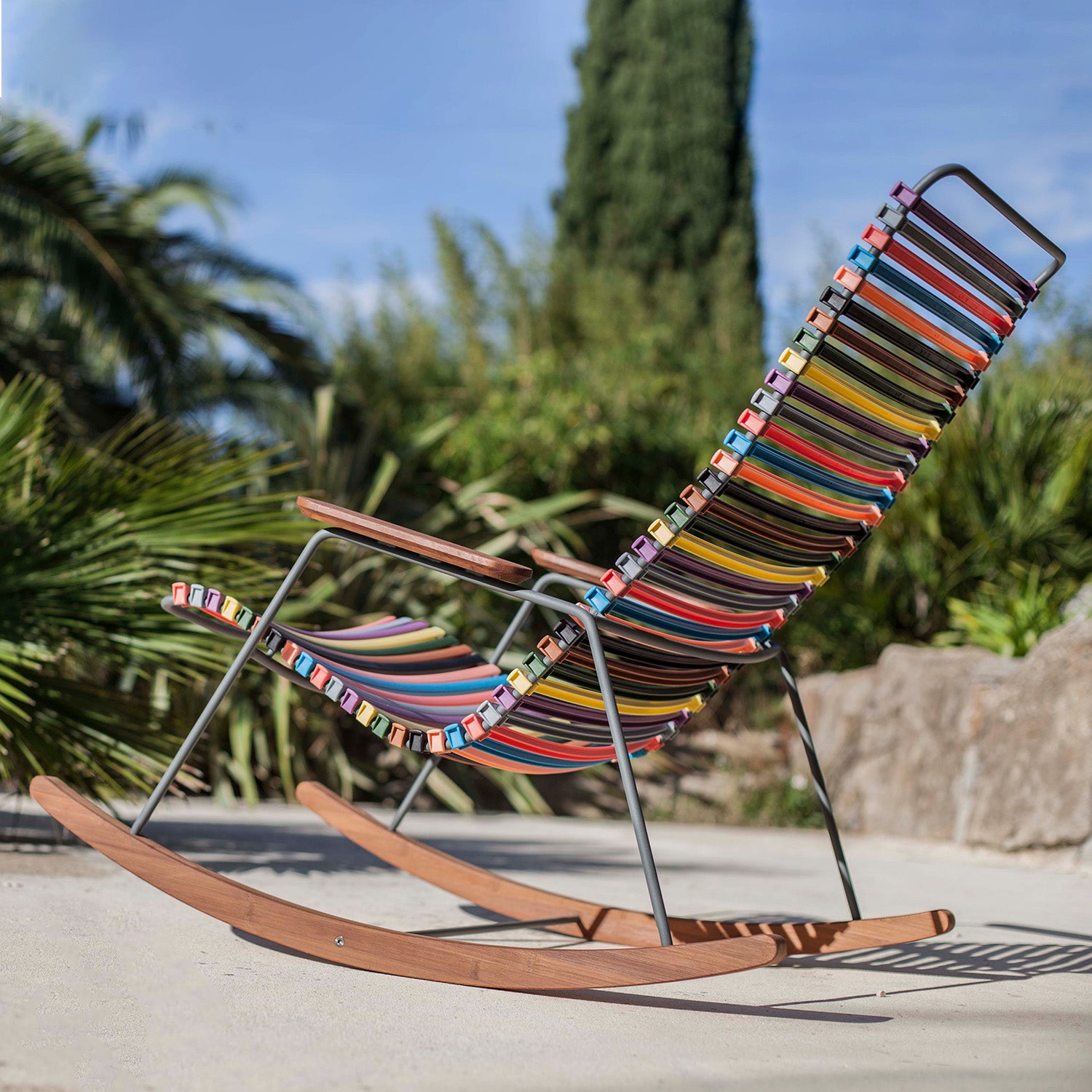The Click Rocking Chair by Houe, featuring a vibrant striped design and a wooden frame adorned with powder-coated steel accents, sits on a sunny patio. Surrounded by lush greenery such as palm leaves and a tall tree, the setting is completed by the blue sky above.