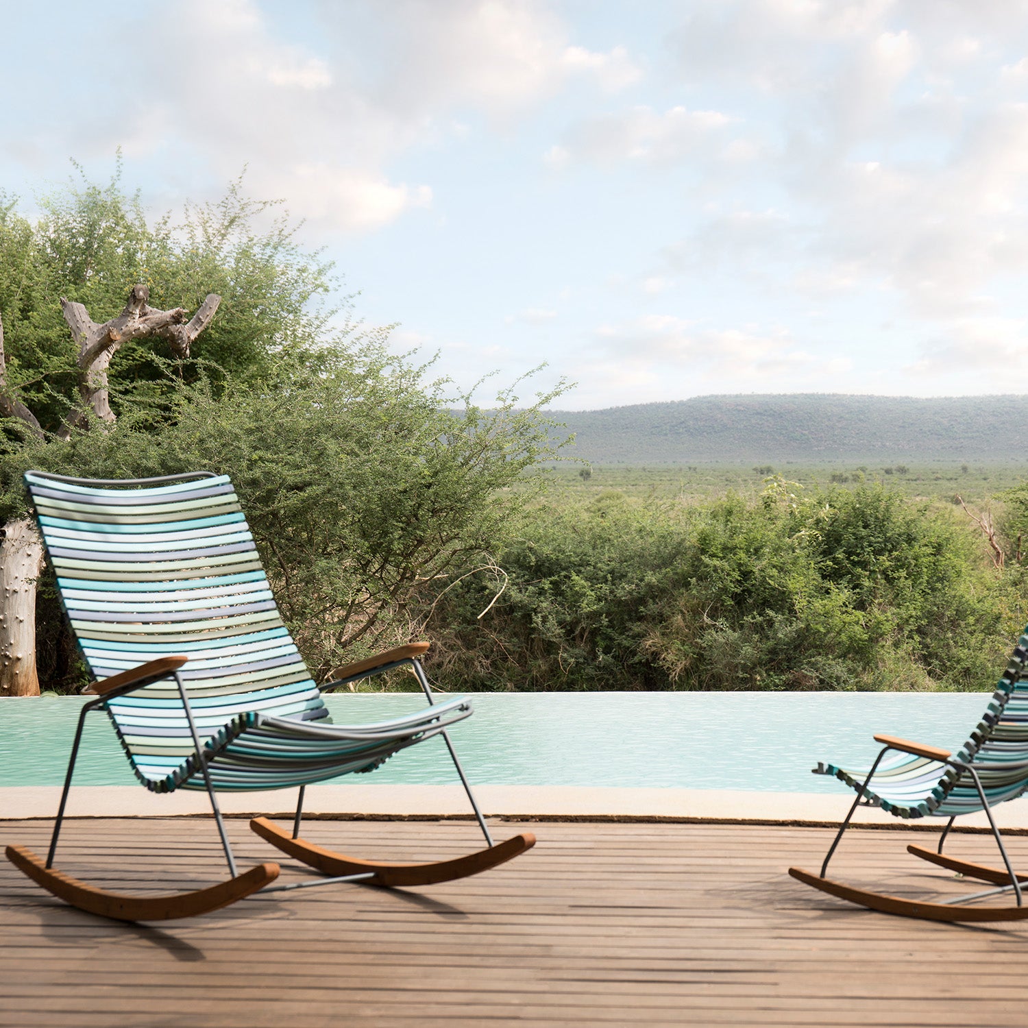 Two Click Rocking Chairs by Houe, made with powder-coated steel and plastic lamellas, adorn a wooden deck beside an infinity pool. Lush greenery stretches behind the pool towards distant hills under a partly cloudy sky, crafting a serene and inviting scene.